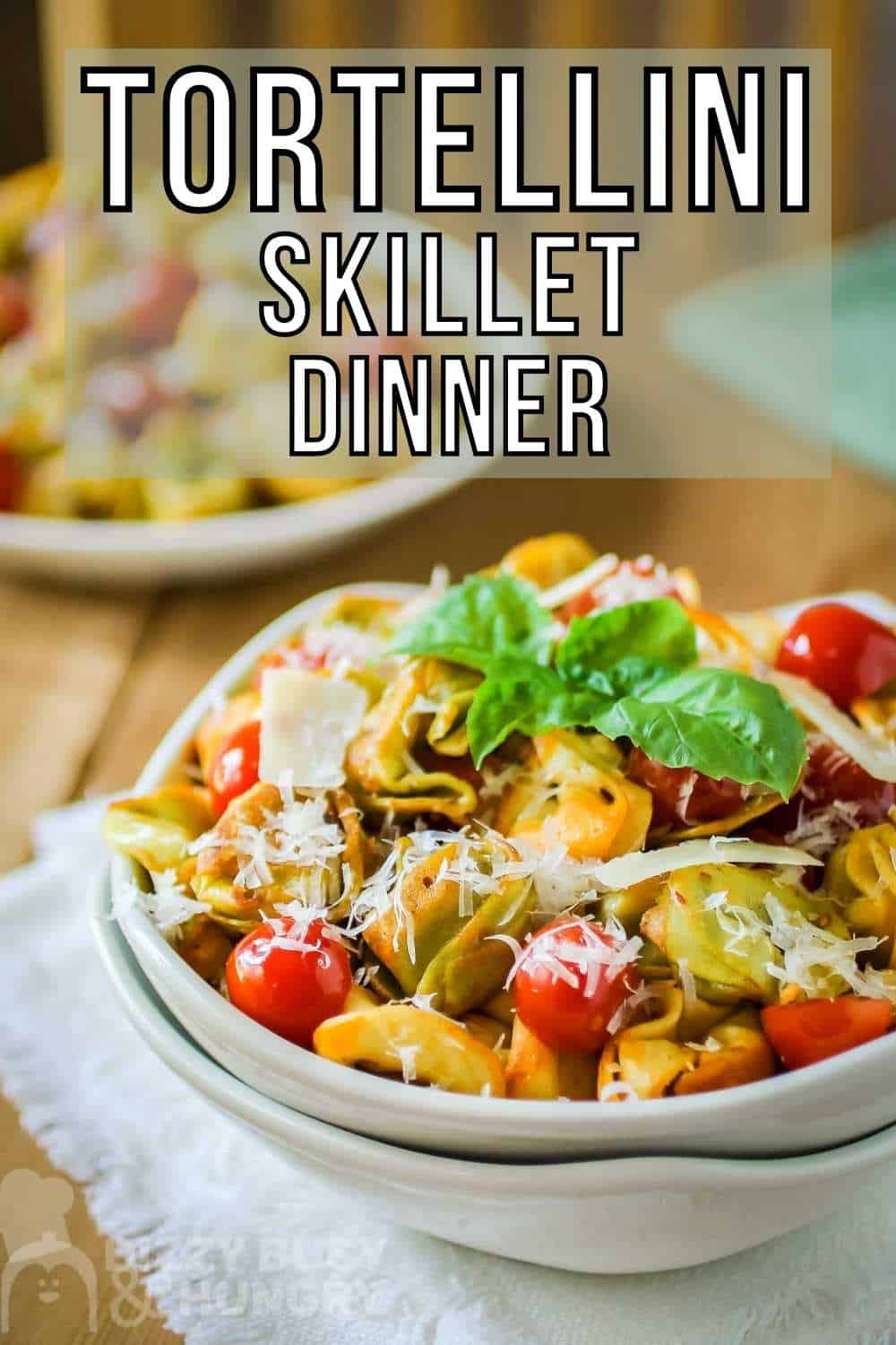 Side angled shot of tortellini garnished with basil and parmesan cheese in a white bowl nestled in another white bowl on a white cloth on a wooden table.