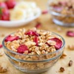 Side view of granola and strawberries in a clear bowl on a wooden surface with more granola and sliced fruits in the background.