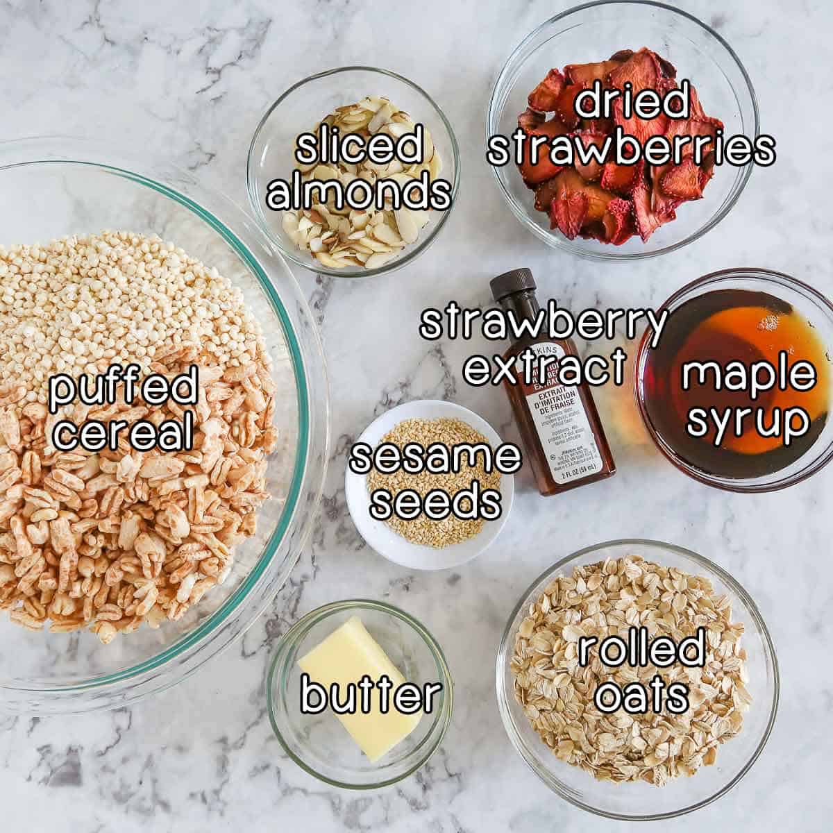 Overhead shot of ingredients- dried strawberries, sliced almonds, puffed cereal, strawberry extract, maple syrup, sesame seeds, butter, and rolled oats.