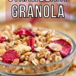 Side view of granola and strawberries in a clear bowl on a wooden surface with more granola in the background.
