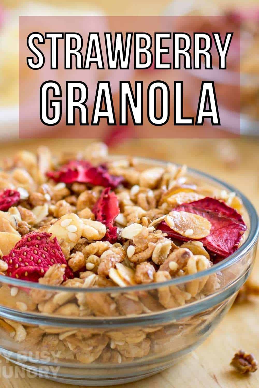 Side view of granola and strawberries in a clear bowl on a wooden surface with more granola in the background.