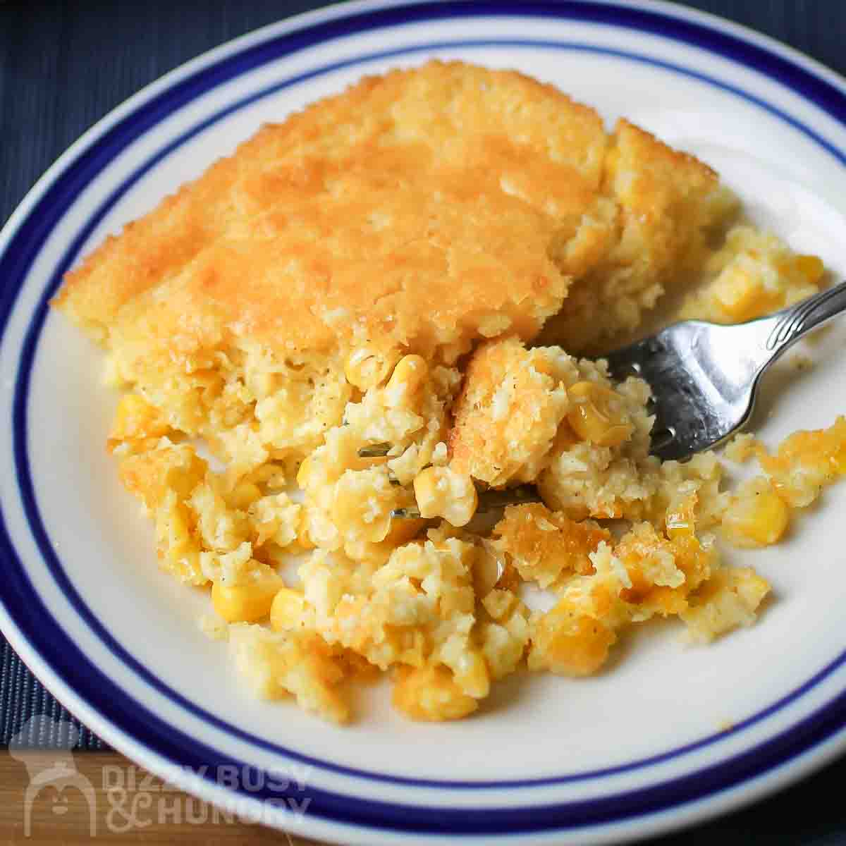 Close up view of corn casserole on a white and blue striped plate with a fork on the side.