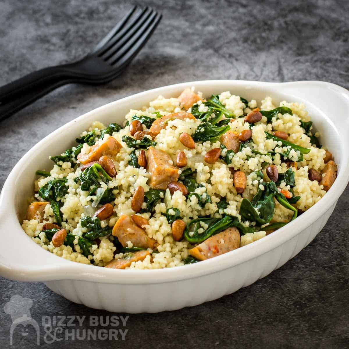 Side angled shot of a white oval dish with couscous and forks in the background on a black and grey surface.