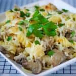 Side shot of turkey Alfredo garnished with herbs on a white square plate on a blue checkered cloth.
