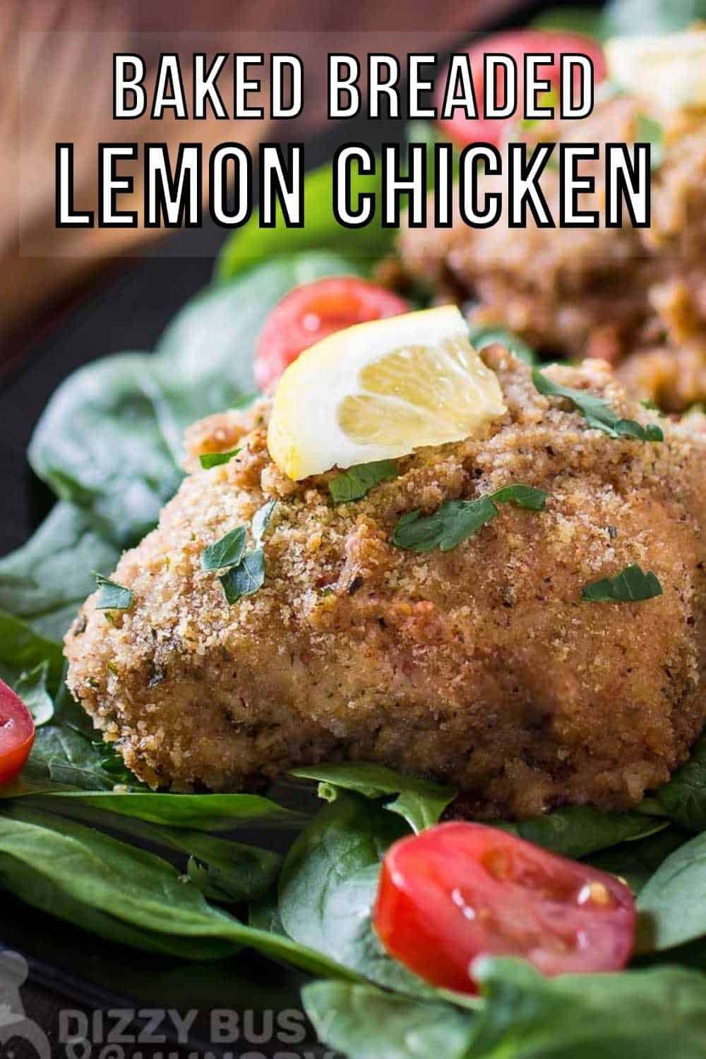 Close up shot of multiple pieces of breaded chicken garnished with herbs and a lemon wedge on a bed on spinach and sliced tomatoes on a black plate.