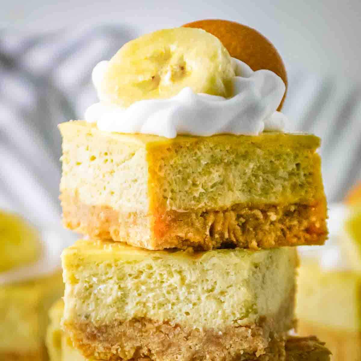 Close up shot of two cheesecake bars stacked on each other garnished with whipped cream, a banana slice, and a vanilla wafer.