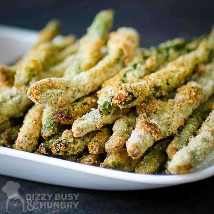 Side view of crispy baked green bean fries stacked on a white plate with a black background.