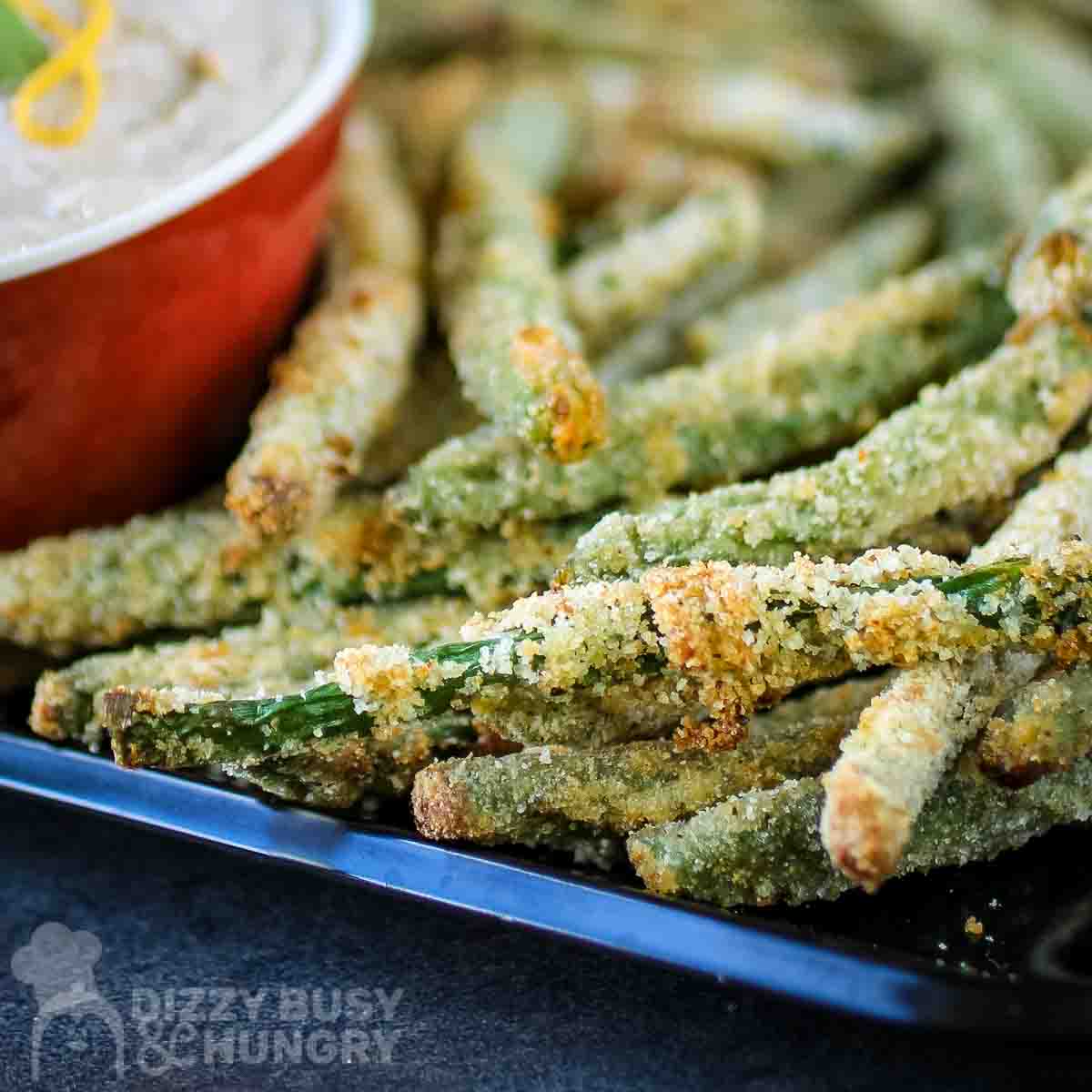 Side close up view of crispy baked green bean fries stacked on a black plate with a red bowl on the side and a black background.