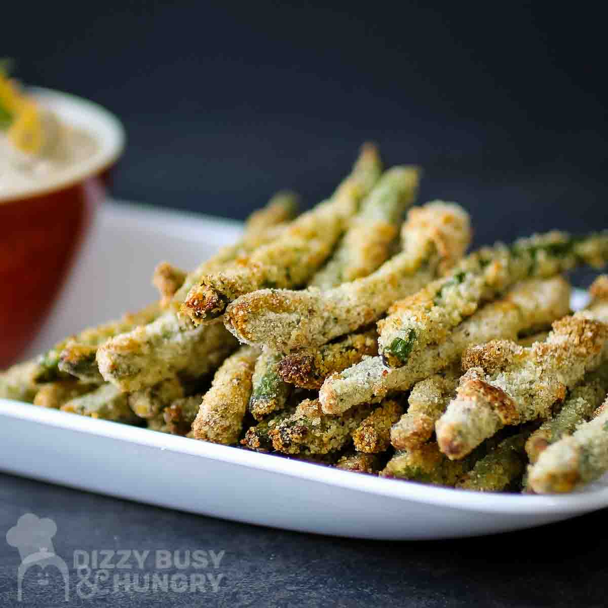 Side view of crispy baked green bean fries stacked on a white plate with a red bowl on the side and a black background.