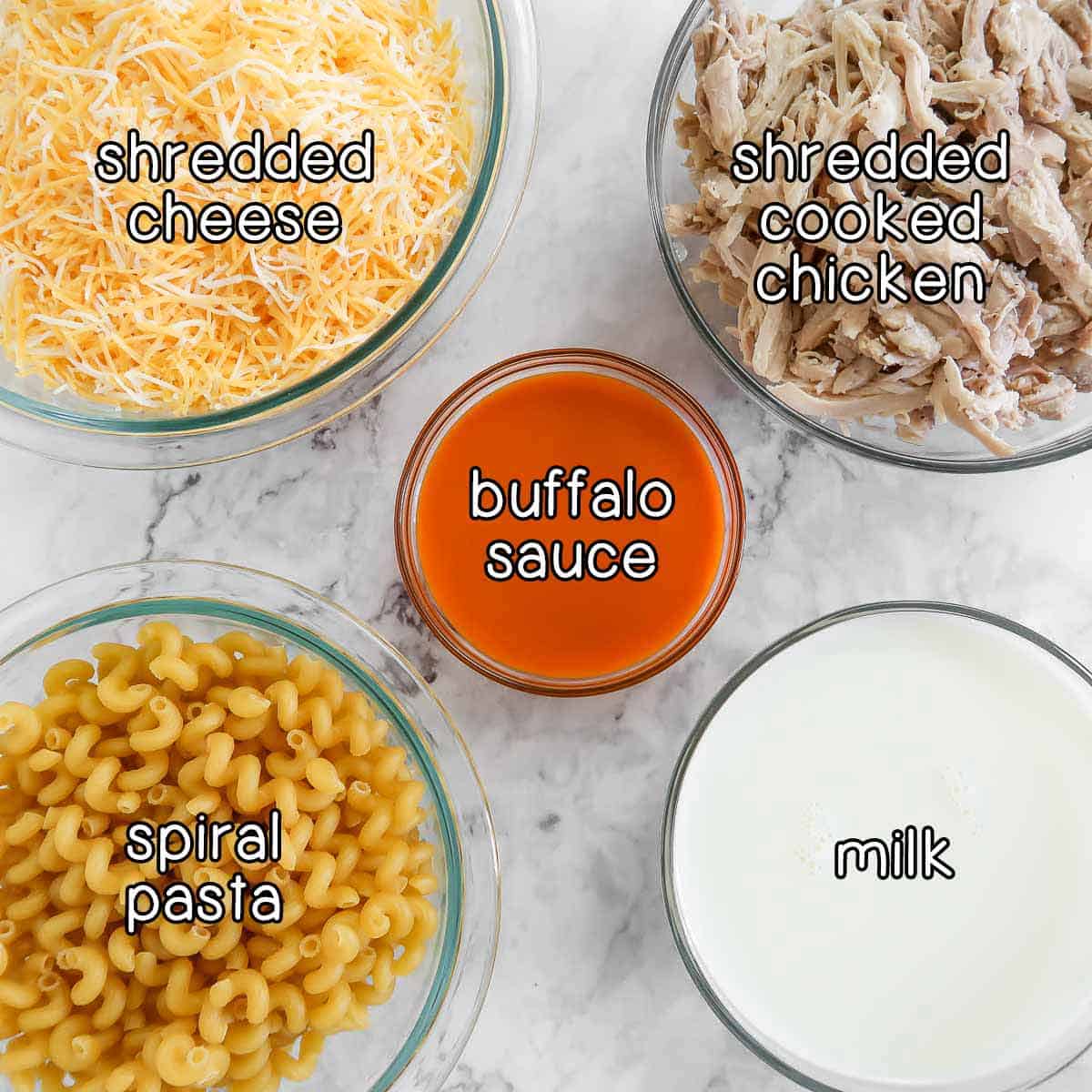 Overhead shot of ingredients - shredded cheese, shredded cooked chicken, buffalo sauce, spiral pasta, and milk.