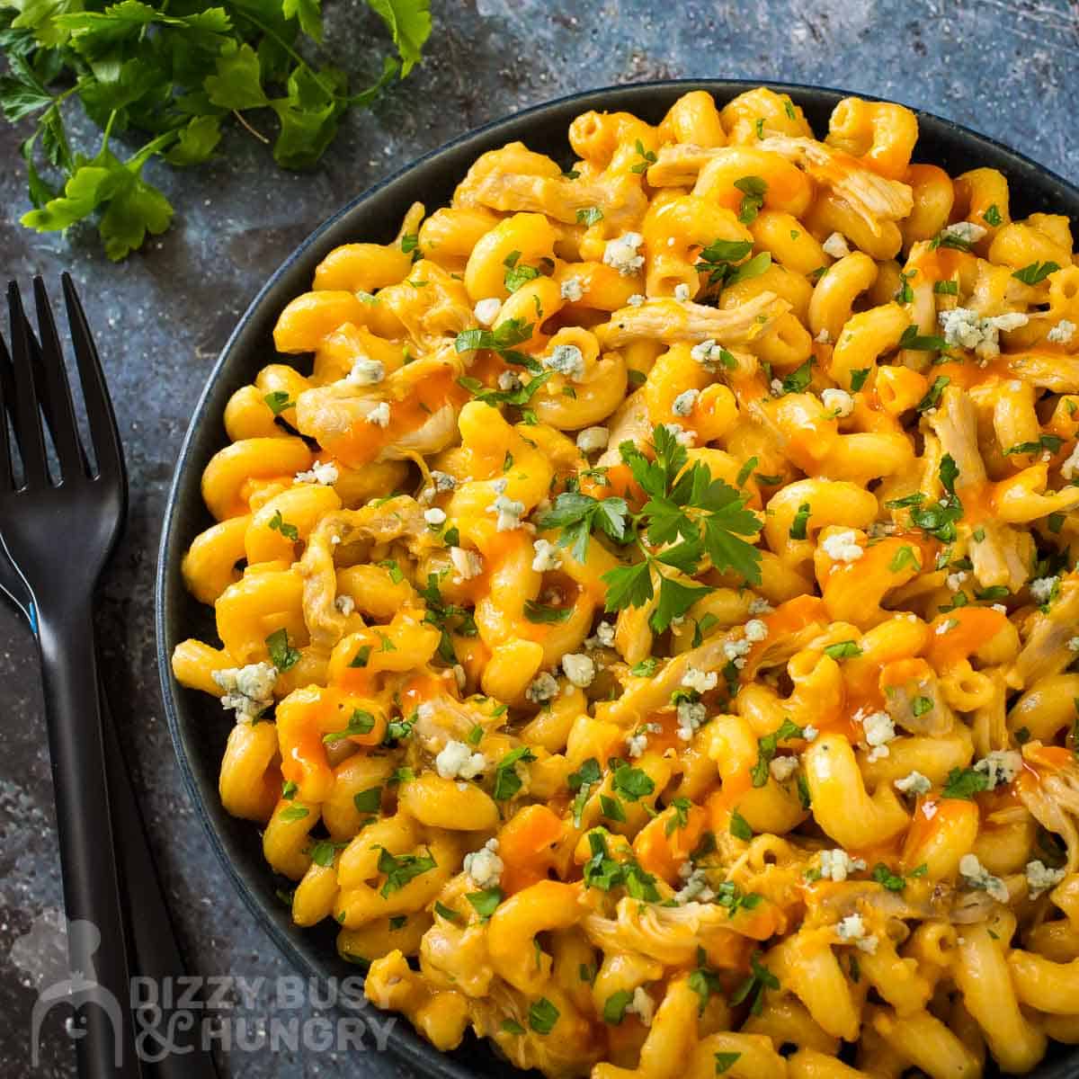 Overhead shot of buffalo Mac and cheese garnished with parsley in a black bowl on a grey and blue marbled surface with a black fork and more parsley in the background.