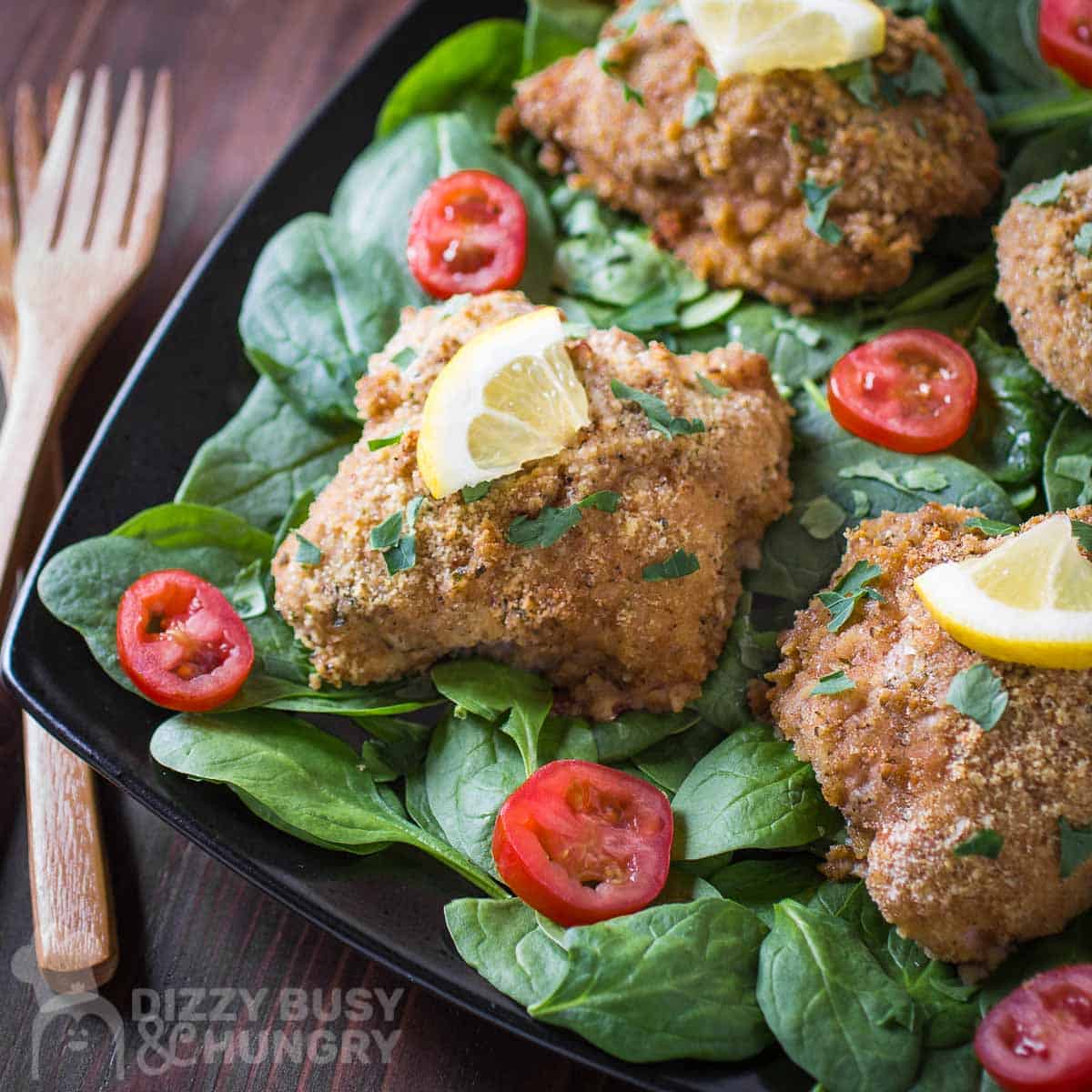 Side shot of multiple pieces of breaded chicken garnished with herbs and a lemon wedge on a bed on spinach and sliced tomatoes on a black plate with wooden forks on the side on a wooden surface.