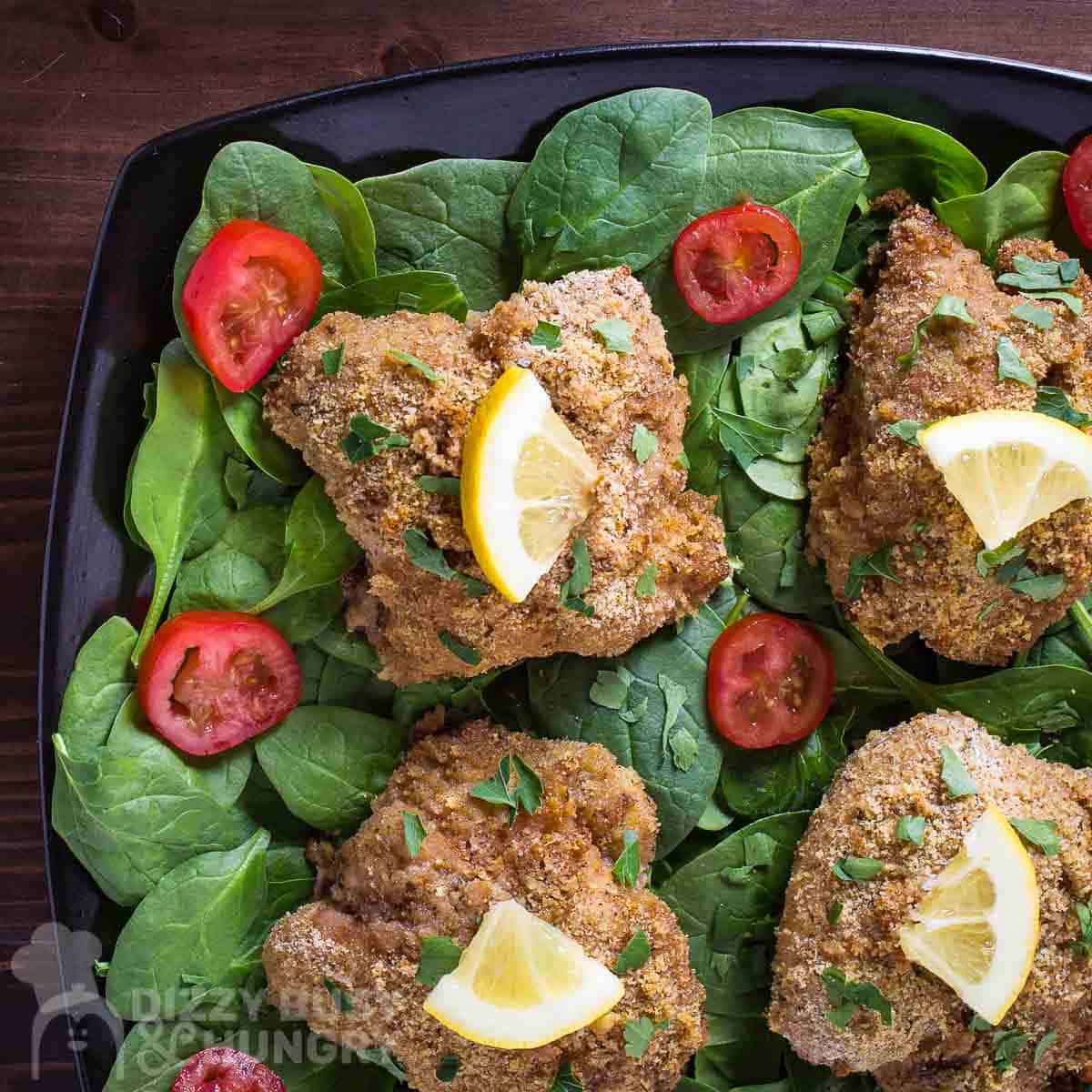 Overhead shot of multiple pieces of breaded chicken garnished with herbs and a lemon wedge on a bed on spinach and sliced tomatoes on a black plate on a wooden table.