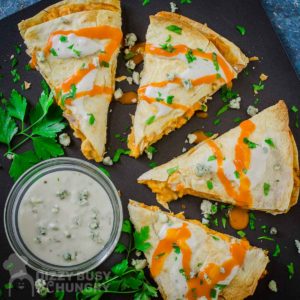 Overhead view of a cut quesadilla drizzled with buffalo sauce and blue cheese garnished with herbs on a black plate with a side of ranch in a white bowl.