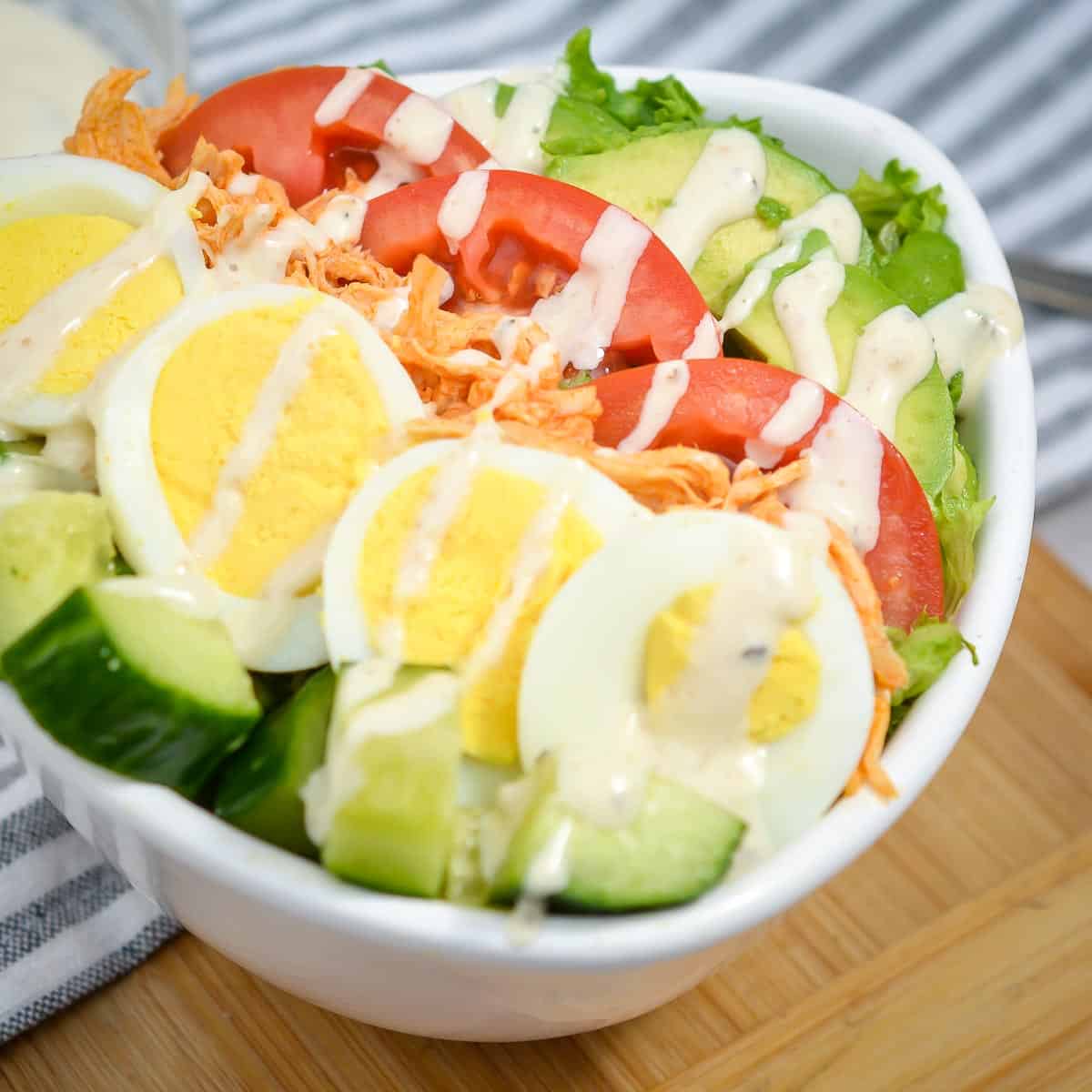 Side close up of buffalo chicken salad with blue cheese dressing in a white bowl on a grey and white striped cloth with a bowl of blue cheese dressing in the background.