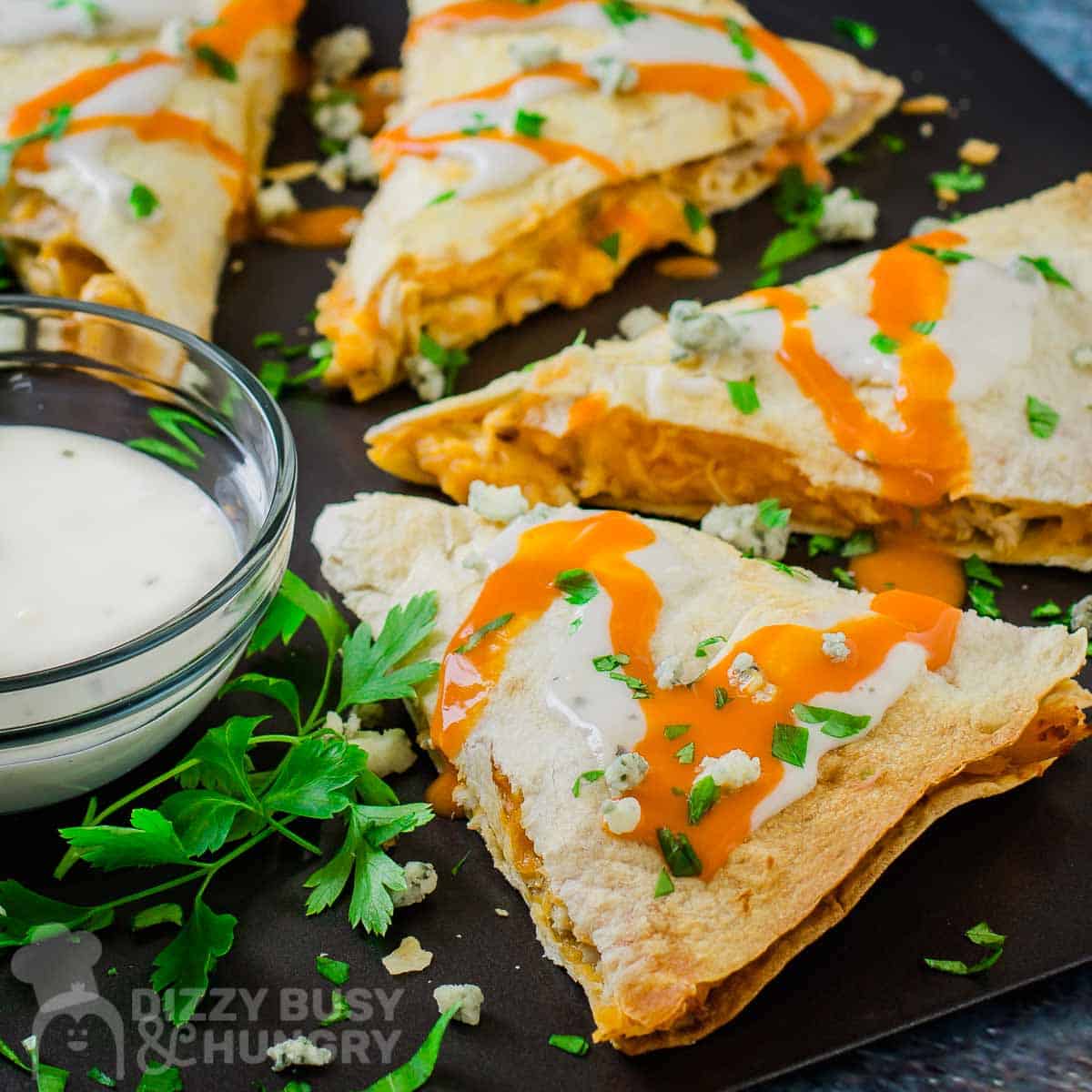 Side angled shot of a cut quesadilla drizzled with buffalo sauce and blue cheese garnished with herbs on a black plate with a side of ranch in a white bowl.