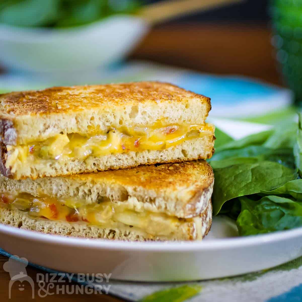 Side shot of two halves of grilled cheese sandwiches stacked on each other on a white plate with a side of greens on a blue and green cloth and a green class in the background.