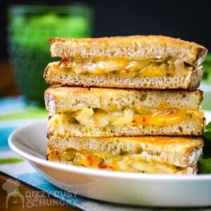 Side shot of three halves of grilled cheese sandwiches stacked on each other on a white plate with a side of greens on a blue and green cloth with a green glass in the background.