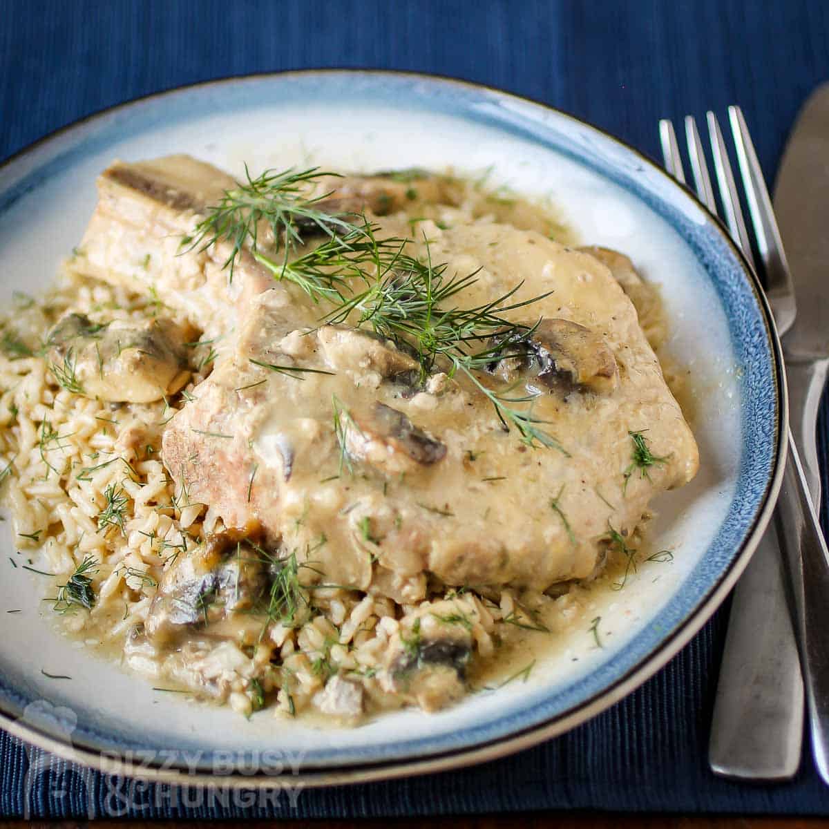 Side close up view of a pork chop on top of mushroom soup garnished with herbs on a blue and white plate with silverware on the side on a dark blue placemat.