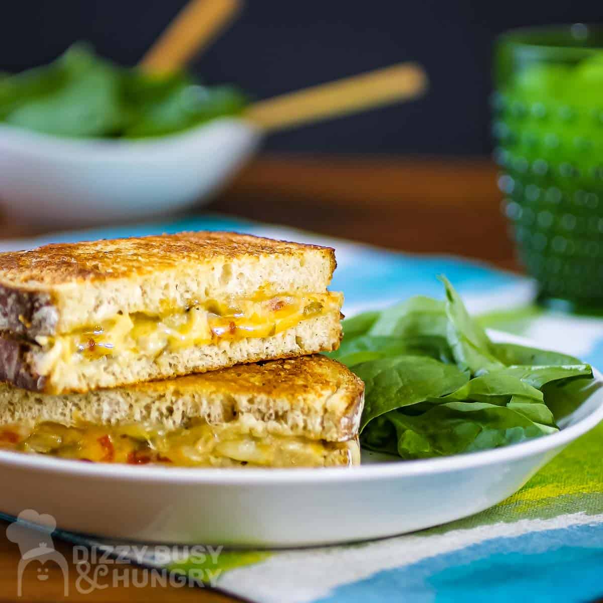 Side shot of two halves of grilled cheese sandwiches stacked on each other on a white plate with a side of greens on a blue and green cloth and a green class in the background.