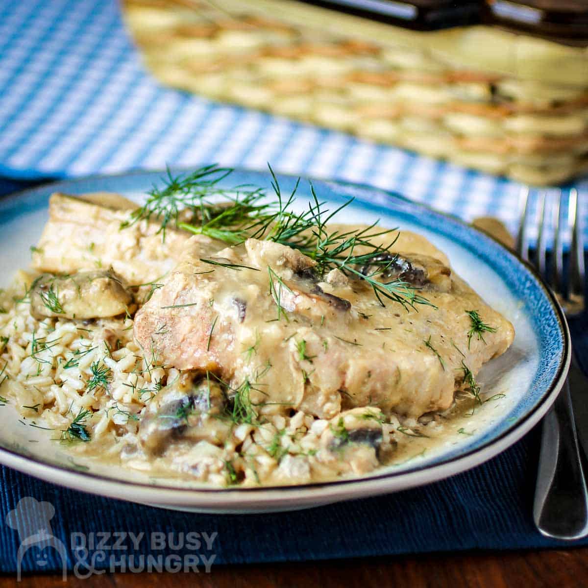 Side angled view of a pork chop on top of mushroom soup garnished with herbs on a blue and white plate with silverware on the side on a dark blue placemat and a basket in the background.