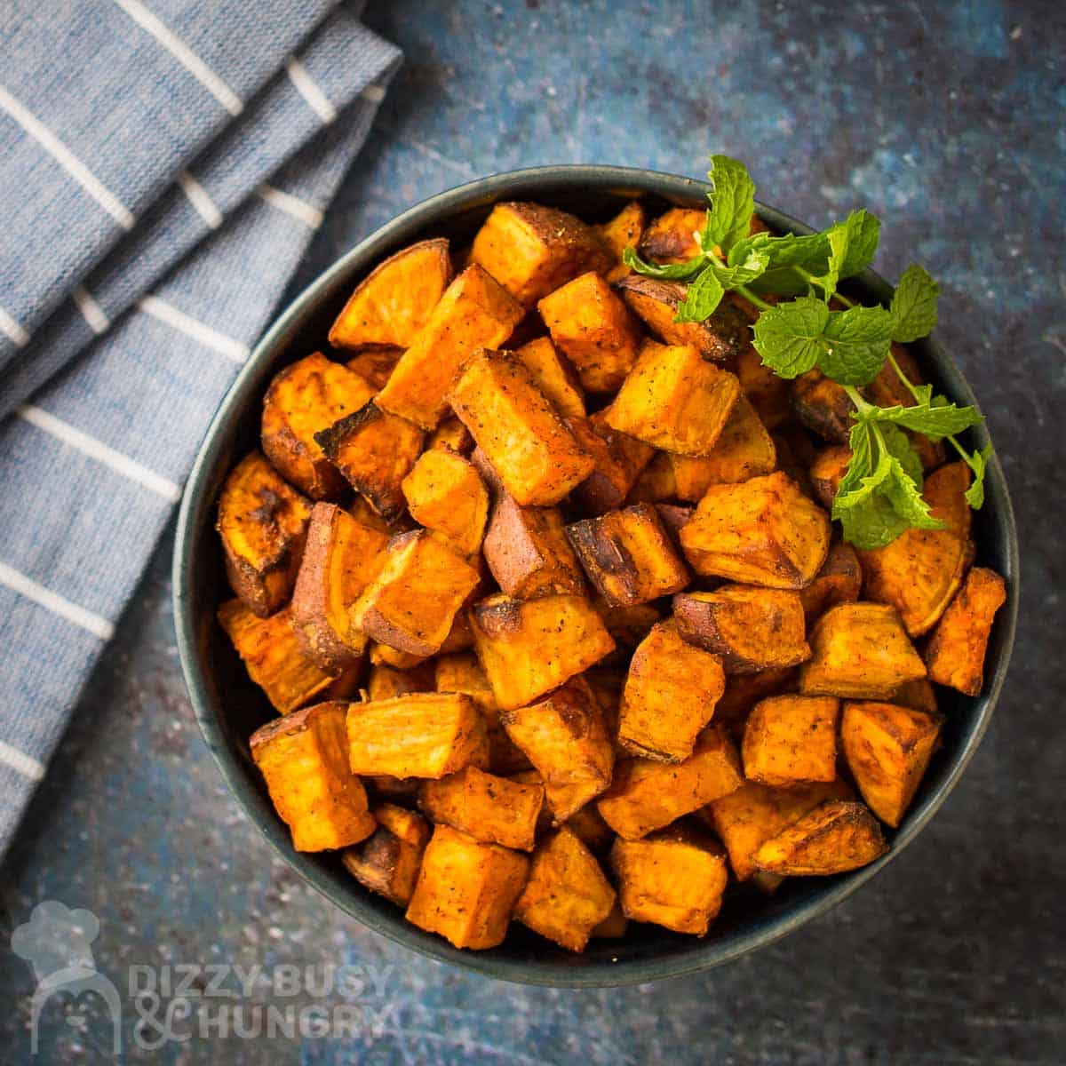 Overhead shot of sweet potato cubes in a black bowl garnished with basil on a blue and grey marble surface with a blue striped towel on the side.