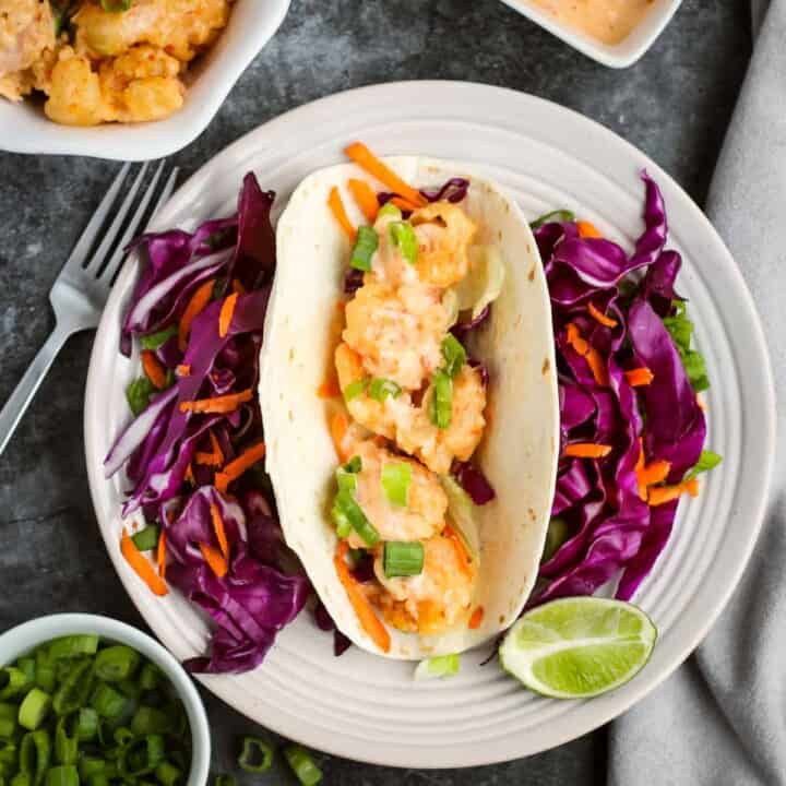 Overhead shot of one shrimp taco with shredded carrots and purple cabbage surrounding it and a lime wedge on the side of the white plate. On the black surface surrounding, there is more shrimp, sauce, and chopped green onions in white bowls as well as a fork and grey cloth.