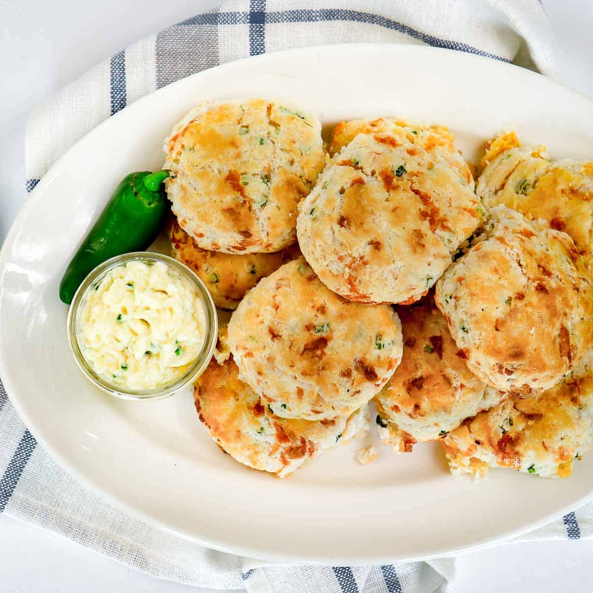 Overhead shot of multiple cheddar biscuits on a large white oval plate with a small glass bowl of jalapeno butter and a full jalapeno on the side all on a blue and white plaid cloth.