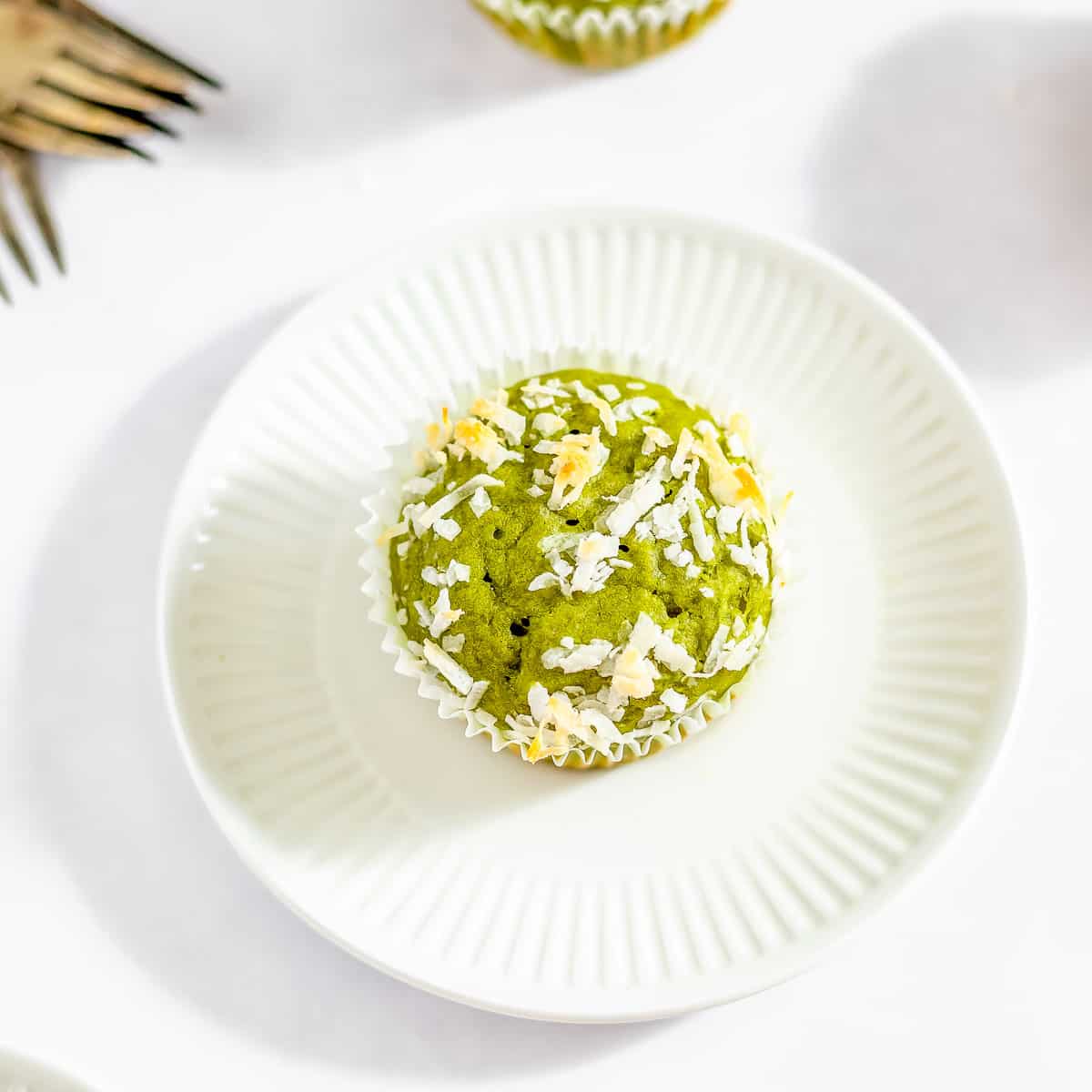 Overhead shot of a matcha muffin garnish with toasted coconut flakes with a white cupcake liner on a white circle plate on a white surface with silverware on the side.