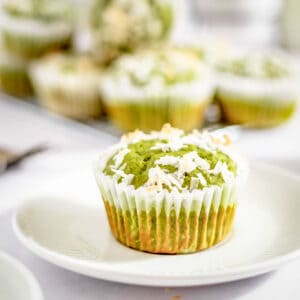 Side shot of one matcha muffin garnished with coconut flakes sitting on a white circle plate with more muffins blurry in the background.