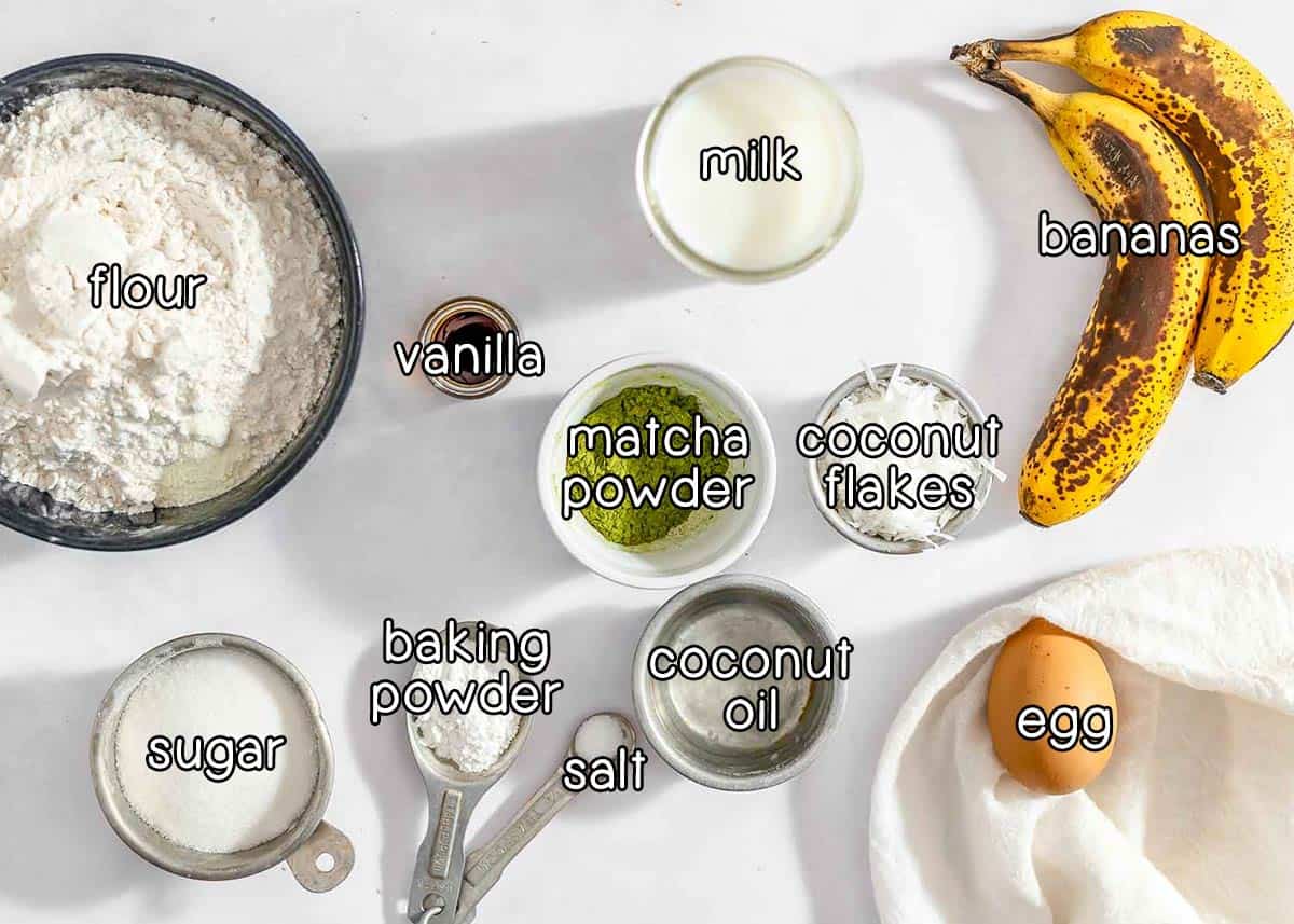 Overhead shot of ingredients - flour, milk, vanilla, matcha powder, coconut flakes, bananas, sugar, baking powder, salt, coconut oil, and an egg.