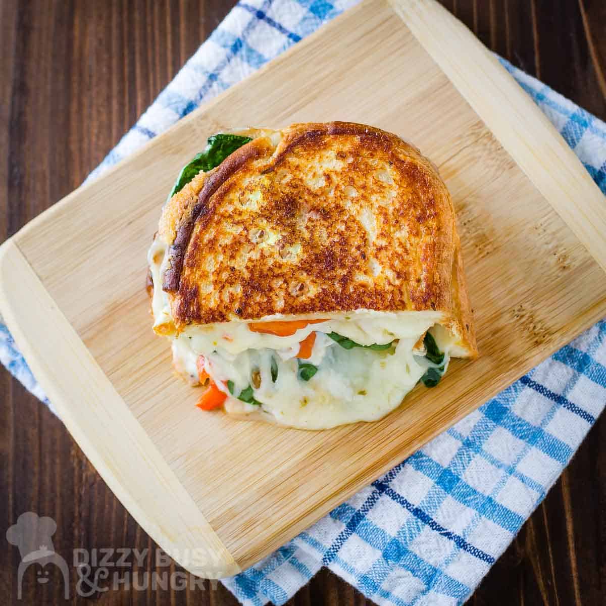 Overhead shot of a grilled cheese split in half with melty cheese on the side on a wooden cutting board on a blue plaid cloth.