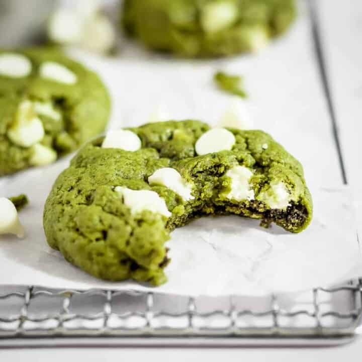 Side shot of a green matcha cookie with a bite taken out on a drying rack with parchment paper with more cookies and white chocolate in the background.