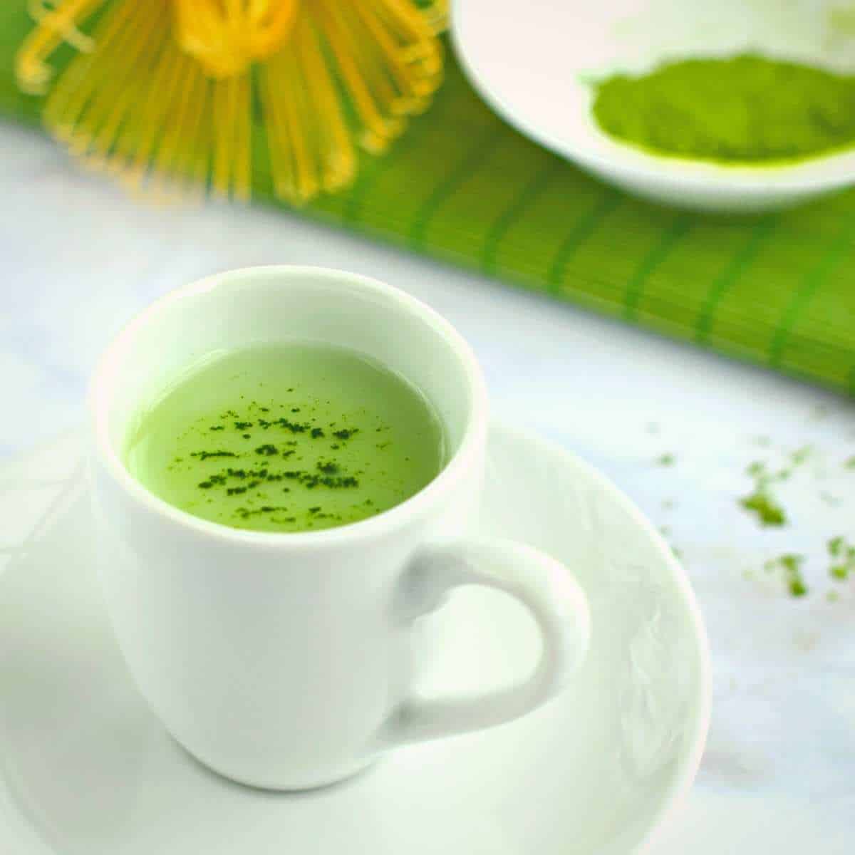 Matcha green tea in a white cup on a white saucer garnished with sprinkled matcha powder on the surface of the tea.