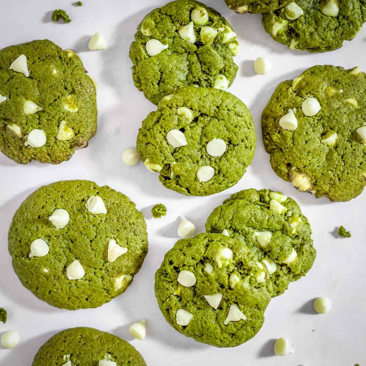 Overhead shot of many matcha cookies scattered on parchment paper with white chocolate chips sprinkled around.