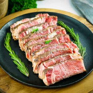 Side angled shot of multiple slices of rare ribeye garnished with salt and rosemary in a row on a black plate on a wooden surface.