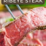 Close up shot of a two pronged fork holding up a slice of rare ribeye with more slices blurry in the background.