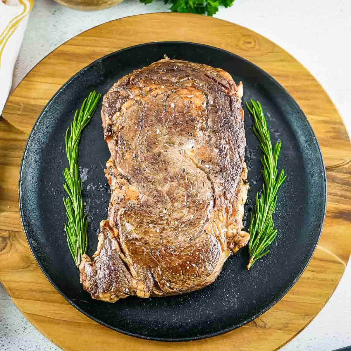 Overhead shot of whole ribeye steak with garnishes of rosemary on the side all on a black plate on a round wooden surface.