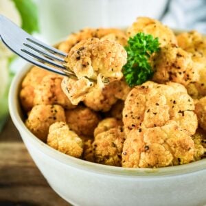 Side shot of a fork picking up a piece of cauliflower from a full cream-colored bowl garnished with fresh herbs.