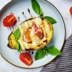 Overhead shot of a baked avocado with tomato basil filling garnished with olive oil, balsamic vinegar, and more basil on a white plate with a black and white striped cloth on the side.