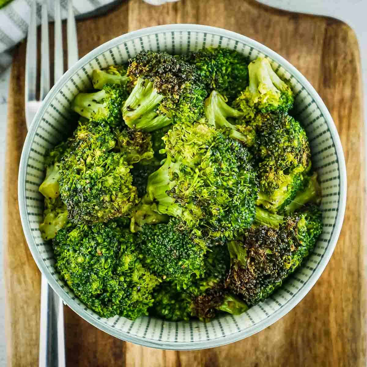 Overhead shot of broccoli in a white and green striped bowl with a fork on the side on a wooden table.