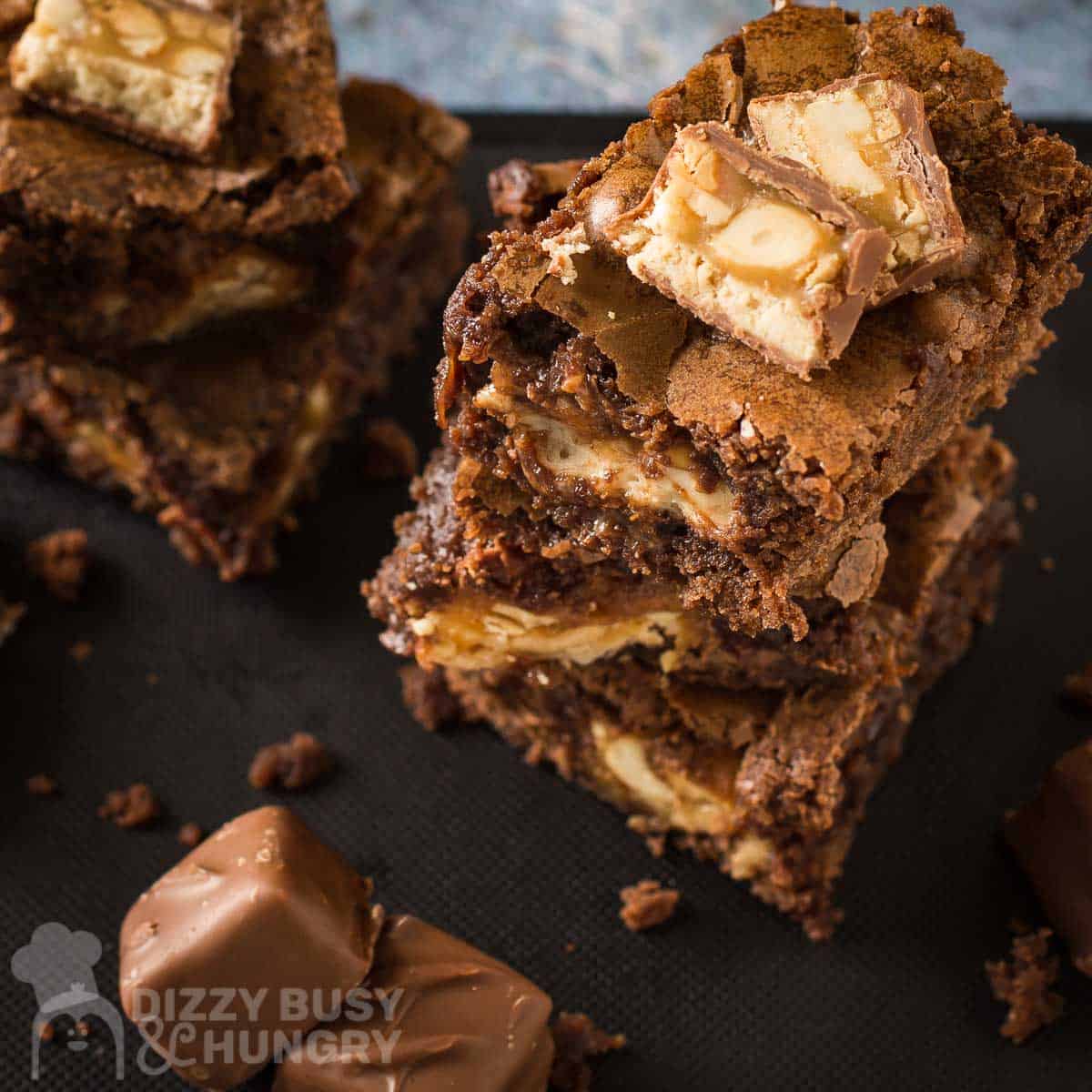 Side shot of multiple snickers brownies stacked on each other with sliced snickers bars on top with full snickers bars on the side all on a black surface.