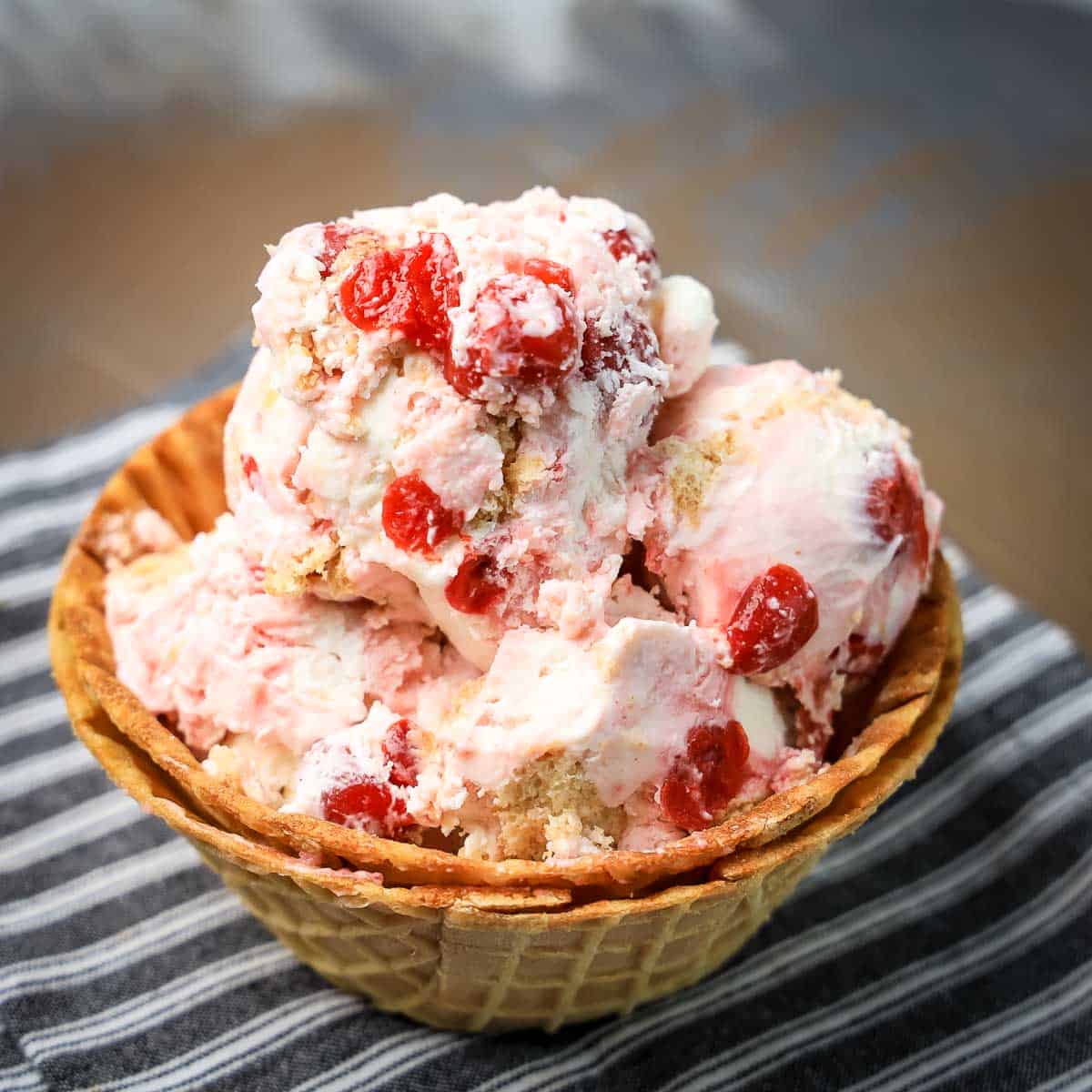 Side angled shot of cherry vanilla ice cream in a waffle cone bowl on a black and white striped cloth.