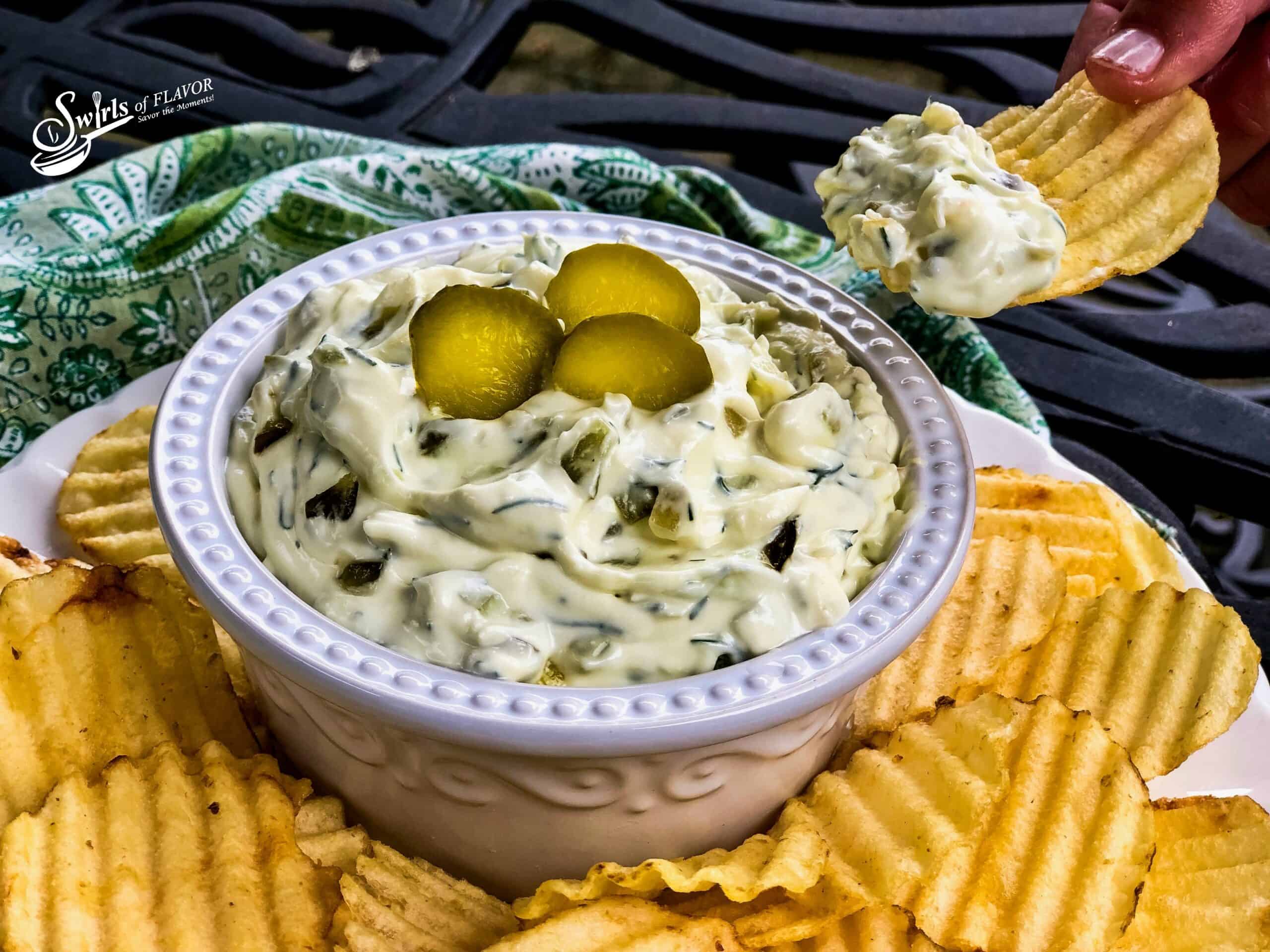 Dill pickle dip in a fancy ceramic bowl topped with 3 dill slices and surrounded by potato chips.