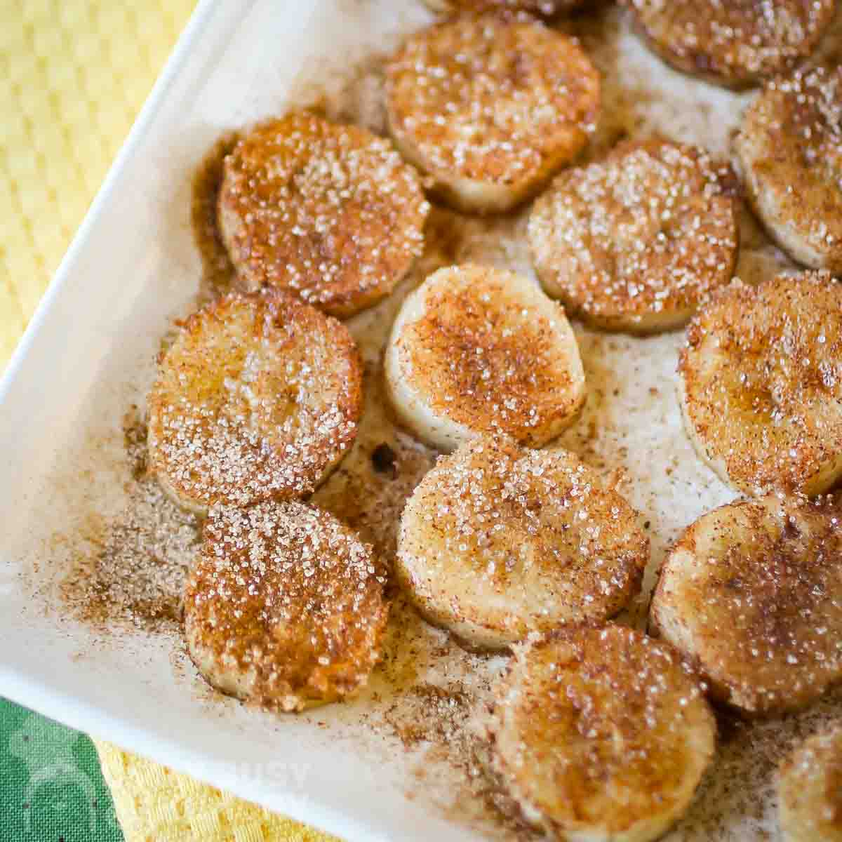 Overhead view of fried banana rounds on a square white dish sitting on a yellow napkin with a green tablecloth.