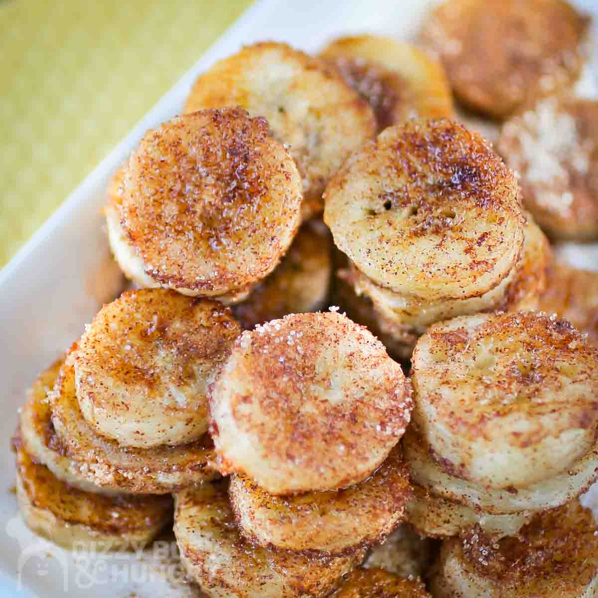 Columns of banana rounds cooked and covered in cinnamon sugar on a white dish placed on a yellow napkin.