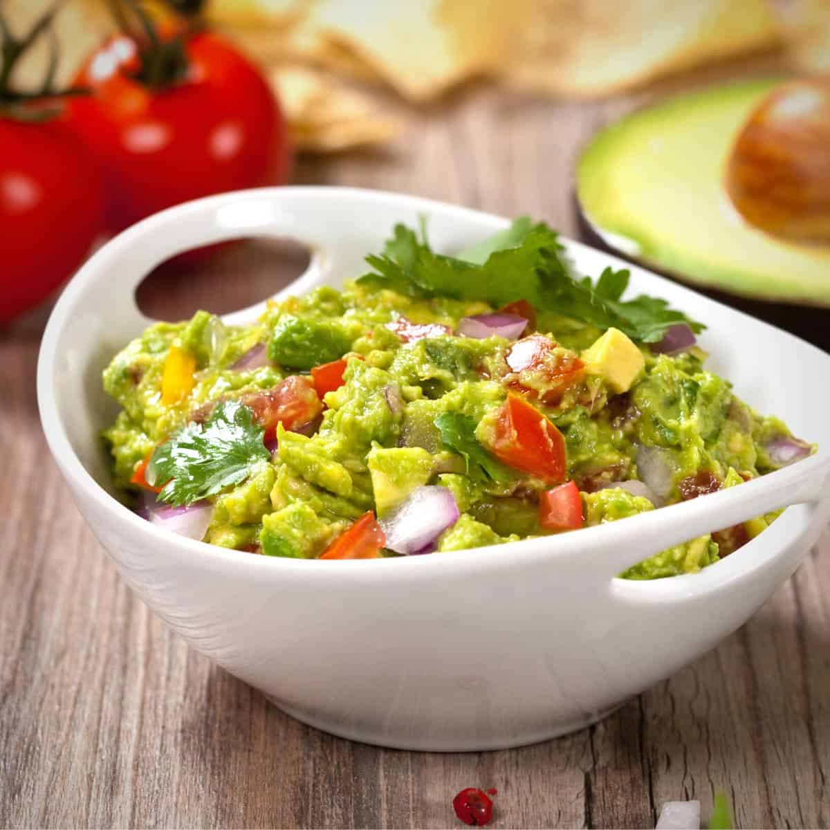 Guacamole in a white bowl on a wooden surface with red tomatoes and an avocado half visible in the background.
