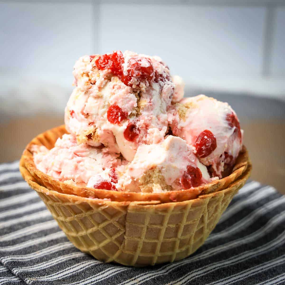 Side shot of cherry vanilla ice cream in a waffle cone bowl on a black and white striped cloth.