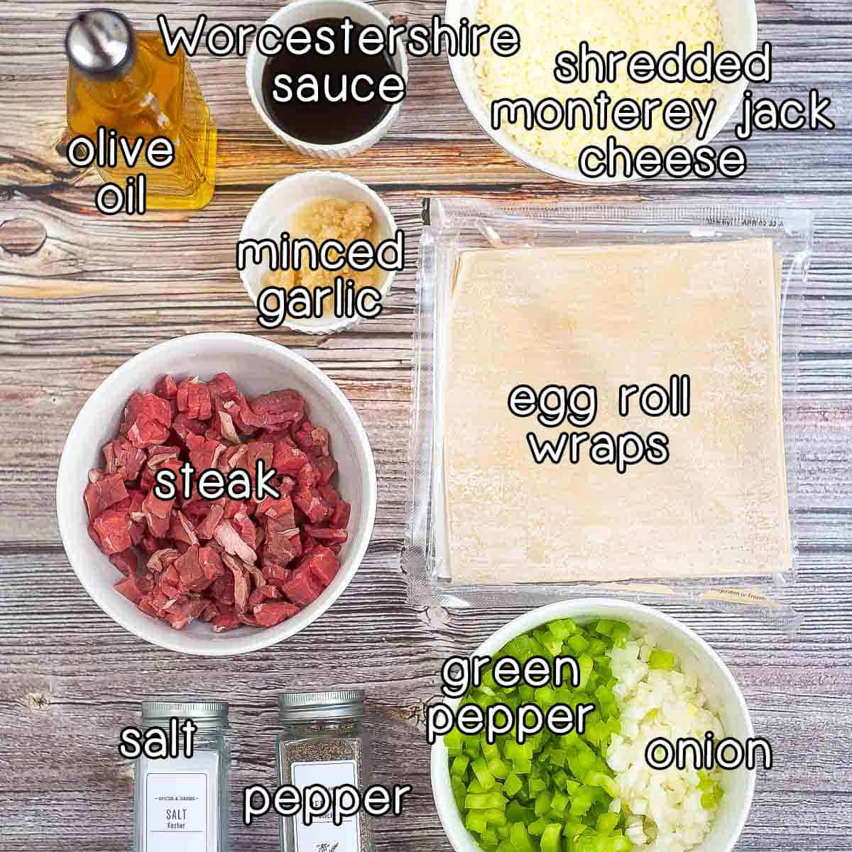 Overhead shot of ingredients - olive oil, Worcestershire sauce, minced garlic, shredded Monterey Jack, steak, egg roll wraps, salt and pepper, green pepper, and onion.