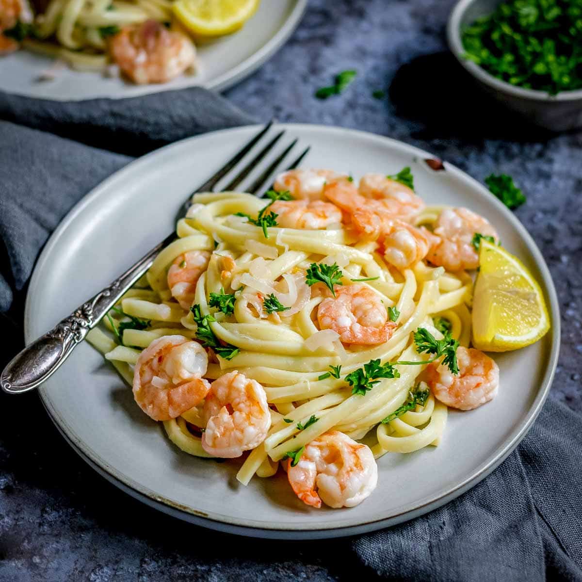 Side shot of shrimp scampi garnished with parsley on a grey plate with a fork on a black and grey surface with a bowl of herbs in the background.